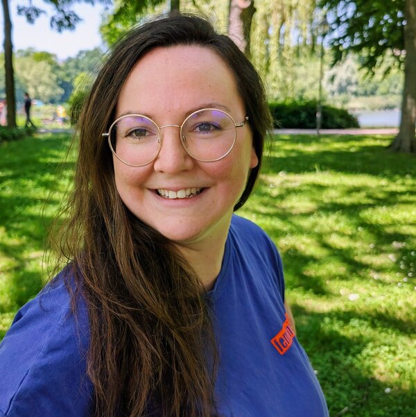 Sina schaut freundlich. Sie hat langes braunes Haar und trägt eine Brille. Ihr T-Shirt ist lila. Sie steht auf einer Wiese.