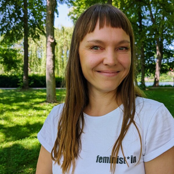 Annett schaut freundlich. Sie hat langes braunes Haar. Ihr T-Shirt ist weiß mit dem Print "feminist*in". Sie steht auf einer Wiese.
