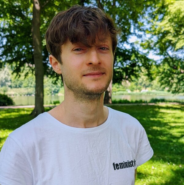 Denny schaut freundlich. Er hat kurzes,gewelltes, braunes Haar. Sein T-Shirt ist weiß mit dem Print "feminist*in". Er steht auf einer Wiese.